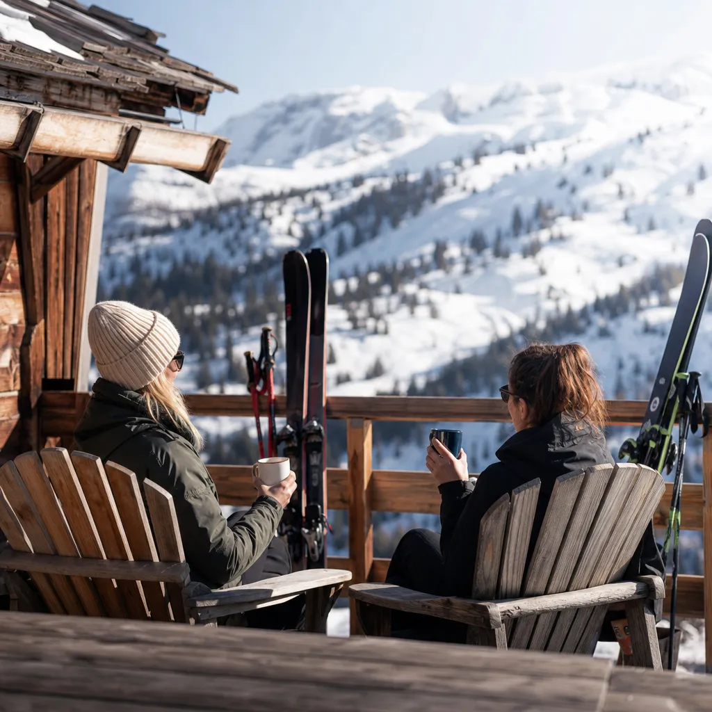 Två vänner i skidkläder tar en fikapaus på en solig terrass vid en alpin skidstuga med skidor lutade mot räcket